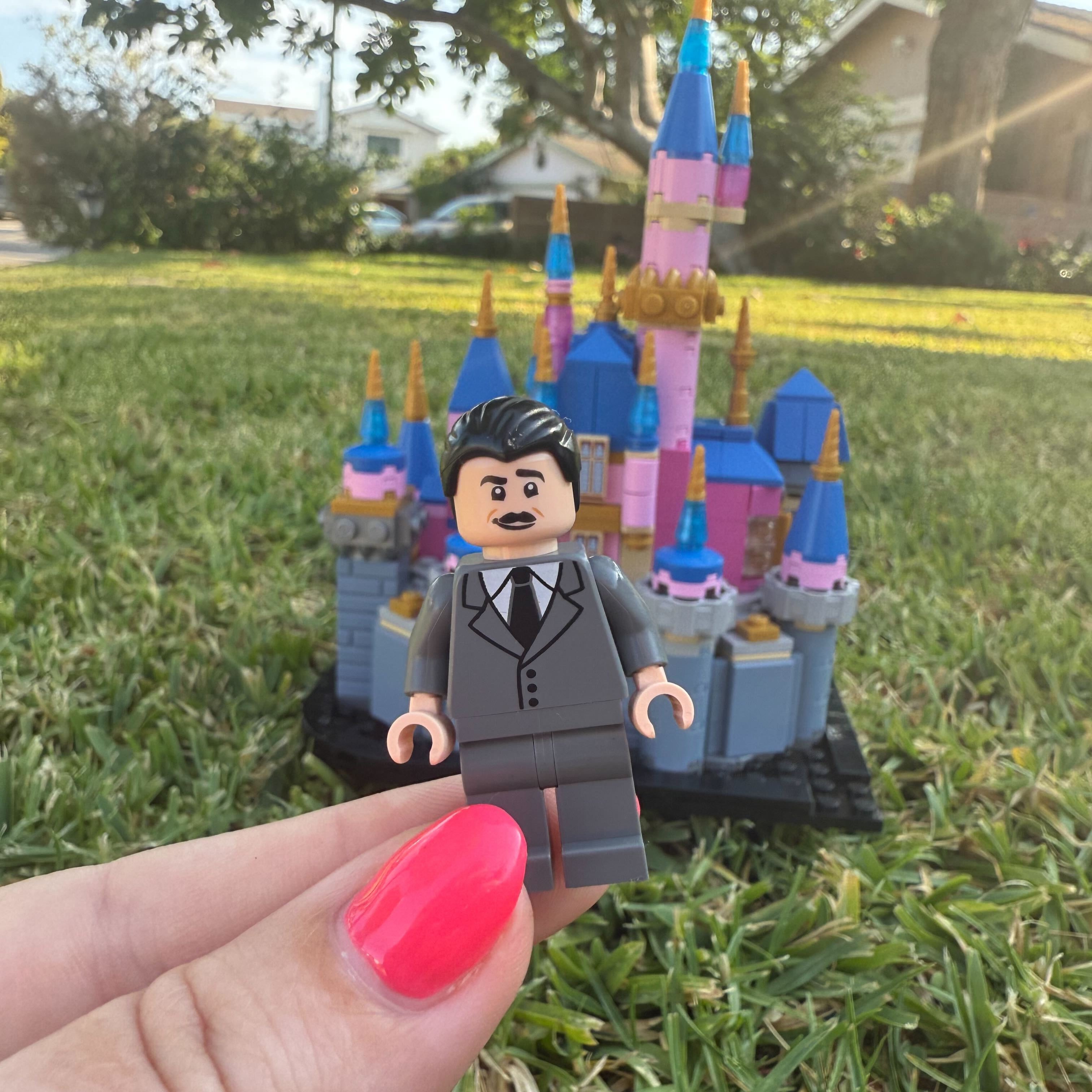 Hand holding a LEGO figure in front of a colorful toy castle on grass